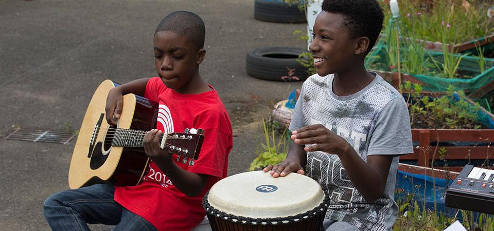 Two young boys making music with a guitar and a drum with plants in containers behind them